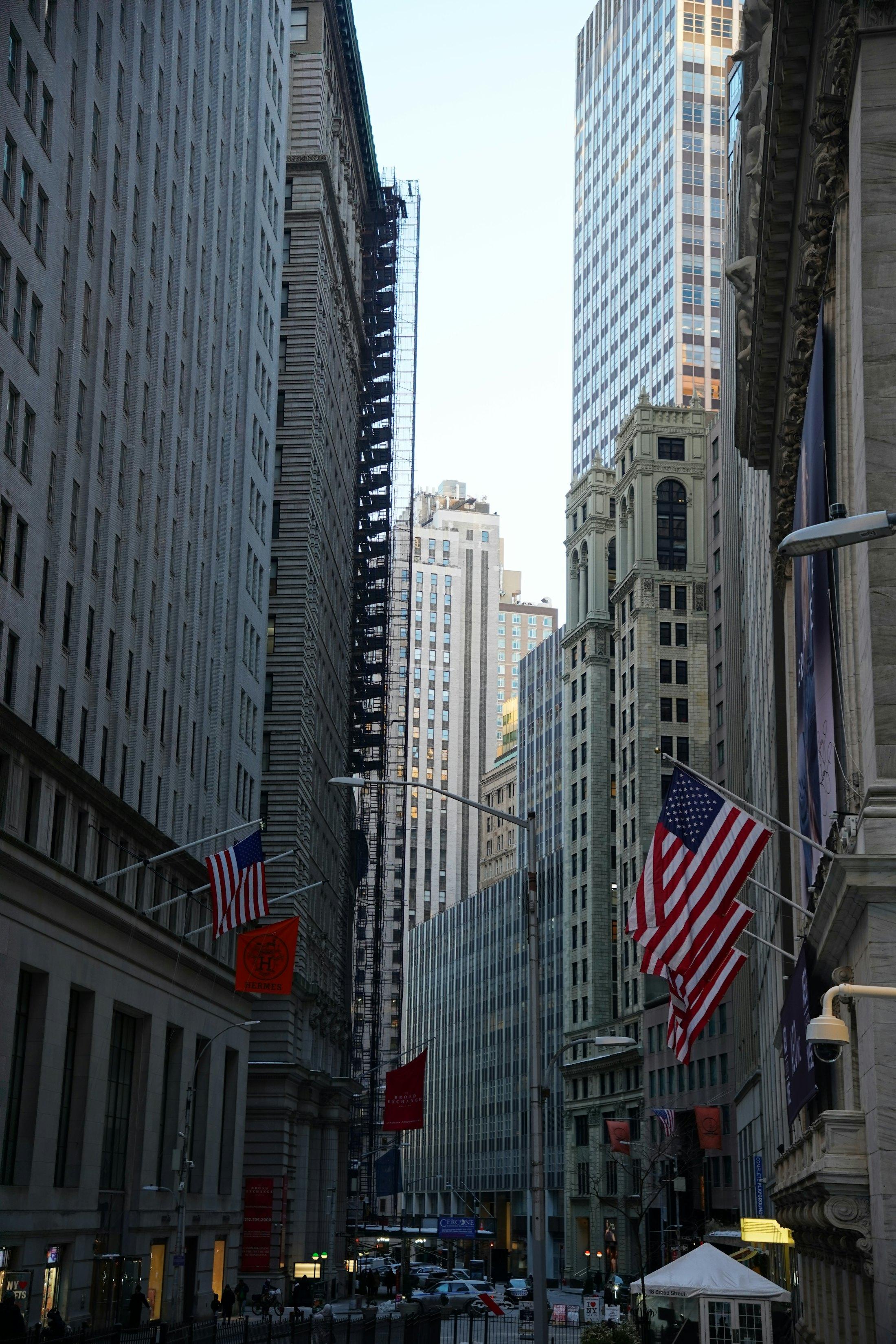 A street of tall buildings in lower Manhattan, flags along it.